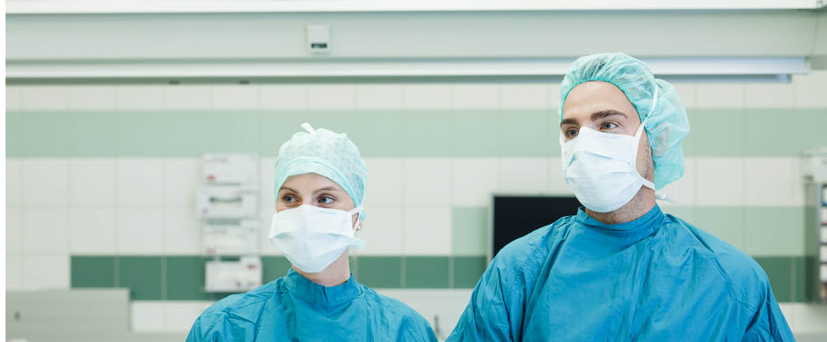 2 Chirurgen in steriler OP-Kleidung, mit Haarnetzen und Mundschutz, stehen am OP-Tisch.